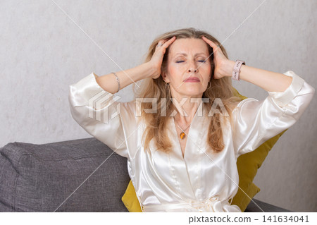 Middle-aged woman relaxing on a couch with eyes closed and hands on her head while wearing a silky robe in a neutral-toned room Middle-aged woman relaxing on a couch with eyes closed and hands on her head while wearing a silky robe in a neutral-toned room 141634041
