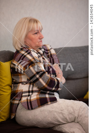 Senior woman sitting on sofa wearing a plaid shirt and light pants, lost in thought 141634045