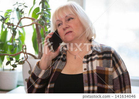 Woman talking on the phone near a potted plant in a bright indoor space Woman talking on the phone near a potted plant in a bright indoor space 141634046