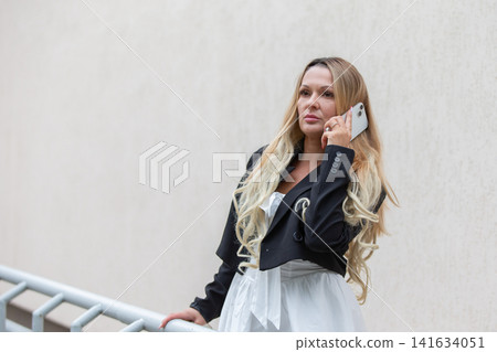 Woman in a black jacket talks on her phone while standing against a light wall Woman in a black jacket talks on her phone while standing against a light wall 141634051