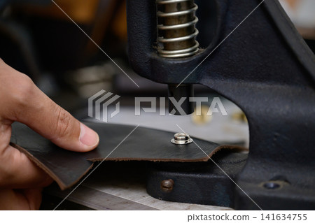 A Japanese man in his 30s using a hand press to attach snaps in leather crafting. A Japanese man in his 30s using a hand press to attach snaps in leather crafting. 141634755