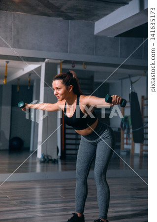 Beauty woman bending forward during focused dumbbell fitness training indoors. Pretty woman exercising upper body muscles with athletic movement in modern gym. Strength wellness concept. Beauty woman bending forward during focused dumbbell fitness training indoors. Pretty woman exercising upper body muscles with athletic movement in modern gym. Strength wellness concept. 141635173
