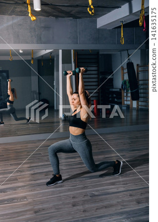 Attractive woman lunging with dumbbells during active gym workout session. Beauty woman exercising lower body muscles with strong athletic posture indoors. Fitness wellness concept. Attractive woman lunging with dumbbells during active gym workout session. Beauty woman exercising lower body muscles with strong athletic posture indoors. Fitness wellness concept. 141635175