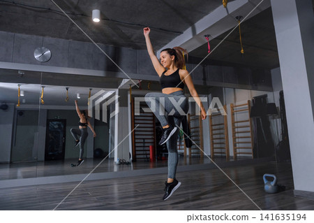 Young woman performing high knee jumps during energetic indoor fitness workout. Beauty woman exercising cardio endurance with athletic balanced posture indoors. Strength wellness concept. Young woman performing high knee jumps during energetic indoor fitness workout. Beauty woman exercising cardio endurance with athletic balanced posture indoors. Strength wellness concept. 141635194