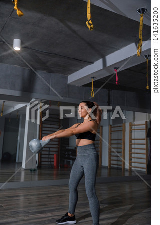 Pretty woman lifting kettlebell during focused indoor strength training routine. Young woman exercising arm muscles with balanced athletic posture inside gym. Fitness wellness concept. Pretty woman lifting kettlebell during focused indoor strength training routine. Young woman exercising arm muscles with balanced athletic posture inside gym. Fitness wellness concept. 141635200