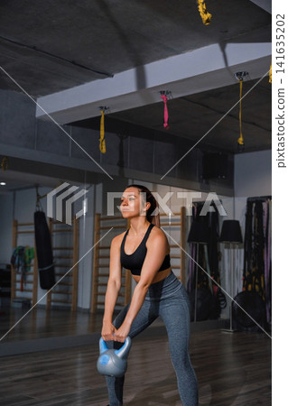 Beauty woman holding kettlebell during focused indoor strength workout session. Attractive woman exercising lower body muscles with balanced athletic posture indoors. Fitness wellness concept. Beauty woman holding kettlebell during focused indoor strength workout session. Attractive woman exercising lower body muscles with balanced athletic posture indoors. Fitness wellness concept. 141635202