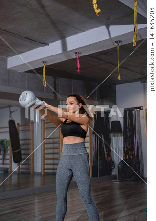 Young woman lifting kettlebell during energetic indoor fitness training routine. Pretty woman exercising upper body muscles with athletic balanced movement inside gym. Strength wellness concept. Young woman lifting kettlebell during energetic indoor fitness training routine. Pretty woman exercising upper body muscles with athletic balanced movement inside gym. Strength wellness concept. 141635203