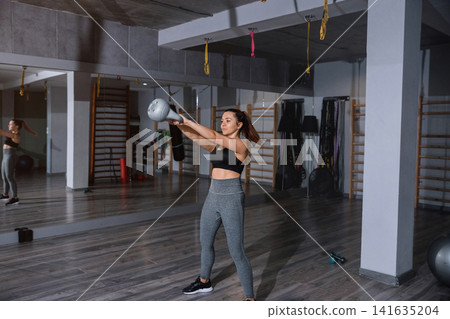 Beautiful woman swinging kettlebell during energetic indoor fitness workout session. Young woman training full body muscles with balanced athletic posture inside gym. Strength wellness concept. Beautiful woman swinging kettlebell during energetic indoor fitness workout session. Young woman training full body muscles with balanced athletic posture inside gym. Strength wellness concept. 141635204