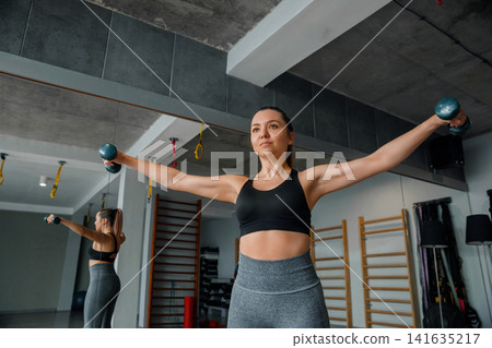 Beauty woman raising dumbbells during energetic indoor shoulder workout routine. Attractive woman exercising upper body muscles with toned athletic movement inside studio. Fitness wellness concept. Beauty woman raising dumbbells during energetic indoor shoulder workout routine. Attractive woman exercising upper body muscles with toned athletic movement inside studio. Fitness wellness concept. 141635217