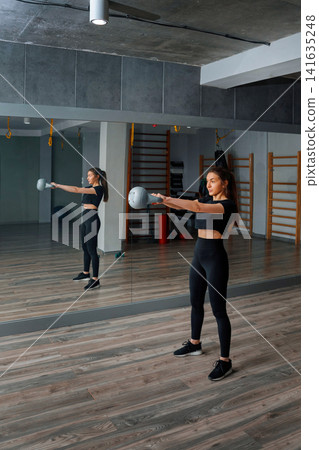 Woman practicing strength training with kettle bell in gym setting during daytime with mirror reflection showing her form and focus on workout Woman practicing strength training with kettle bell in gym setting during daytime with mirror reflection showing her form and focus on workout 141635248