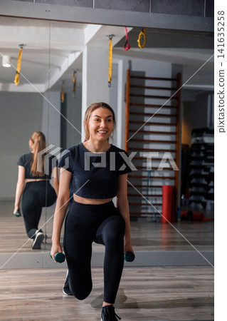 Beauty woman performing lunges with dumbbells in bright gym. Attractive woman smiling during lower body fitness training routine indoors. Wellness workout concept. Beauty woman performing lunges with dumbbells in bright gym. Attractive woman smiling during lower body fitness training routine indoors. Wellness workout concept. 141635258