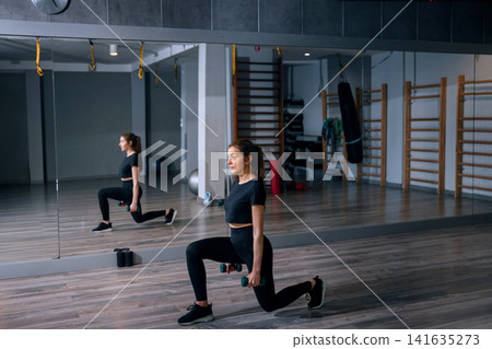 Beautiful woman performing lunges during active fitness gym workout. Young woman training lower body muscles with focused athletic posture indoors. Strength wellness concept. Beautiful woman performing lunges during active fitness gym workout. Young woman training lower body muscles with focused athletic posture indoors. Strength wellness concept. 141635273