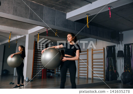 Beautiful woman holding fitness ball inside modern workout gym studio. Young woman preparing active exercise routine with healthy athletic posture indoors. Wellness sport concept. Beautiful woman holding fitness ball inside modern workout gym studio. Young woman preparing active exercise routine with healthy athletic posture indoors. Wellness sport concept. 141635278