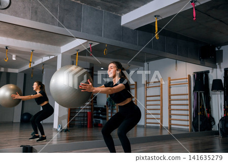Beauty woman exercising with stability ball during active fitness training. Attractive woman performing squats with athletic posture inside modern gym studio. Strength wellness concept. Beauty woman exercising with stability ball during active fitness training. Attractive woman performing squats with athletic posture inside modern gym studio. Strength wellness concept. 141635279
