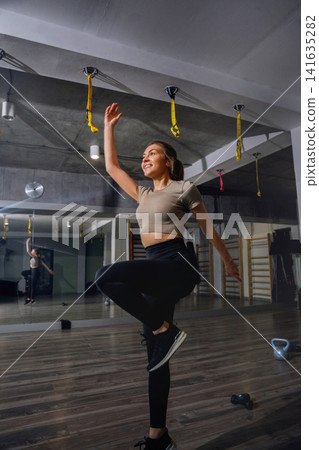 Woman practices fitness routine in gym area with mirrors and exercise equipment during daytime Woman practices fitness routine in gym area with mirrors and exercise equipment during daytime 141635282