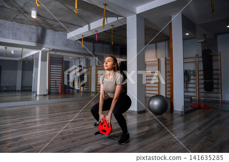 Woman performs squat exercise with kettlebell at gym during daytime workout session Woman performs squat exercise with kettlebell at gym during daytime workout session 141635285