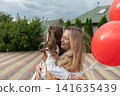 Mother and daughter enjoy a warm embrace while holding red balloons in a garden during a cloudy afternoon 141635439