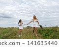 Two girls stroll hand in hand through a sunny field while wearing white dresses and holding wildflowers 141635440