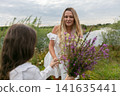 Woman in white dress receives flowers from child near serene river and greenery during overcast day 141635441