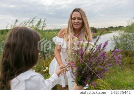 Woman in white dress receives flowers from child near serene river and greenery during overcast day Woman in white dress receives flowers from child near serene river and greenery during overcast day 141635441