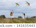 Two girls fly colorful kites in a grassy field under a cloudy sky during a breezy afternoon 141635443
