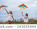 Two girls flying colorful kites in an open field on a cloudy day with soft grasses surrounding them 141635444