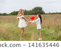 Two girls in white dresses hold hands in a grassy field while playing with a red balloon on a cloudy day 141635446