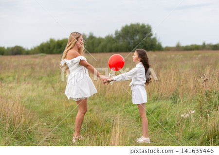 Two girls in white dresses hold hands in a grassy field while playing with a red balloon on a cloudy day Two girls in white dresses hold hands in a grassy field while playing with a red balloon on a cloudy day 141635446