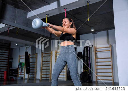 Fitness enthusiast swings a kettlebell in a gym during the day focused on strength training and improving physical health Fitness enthusiast swings a kettlebell in a gym during the day focused on strength training and improving physical health 141635523