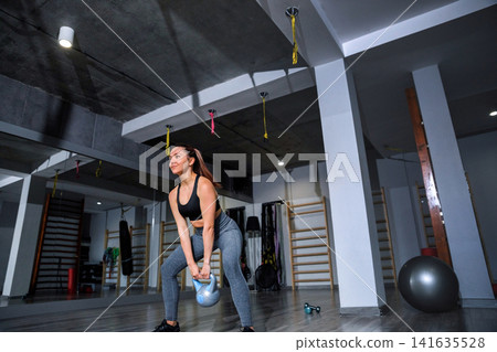 Woman lifts kettlebell in modern gym during workout session focused on strength training and fitness routine Woman lifts kettlebell in modern gym during workout session focused on strength training and fitness routine 141635528