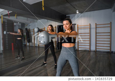 Two women workout together in a gym using kettlebells, engaging in strength training in a mirrored space with fitness equipment Two women workout together in a gym using kettlebells, engaging in strength training in a mirrored space with fitness equipment 141635729