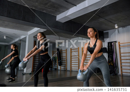 Two women exercise with kettlebells in a gym studio during a group training session in the afternoon Two women exercise with kettlebells in a gym studio during a group training session in the afternoon 141635731