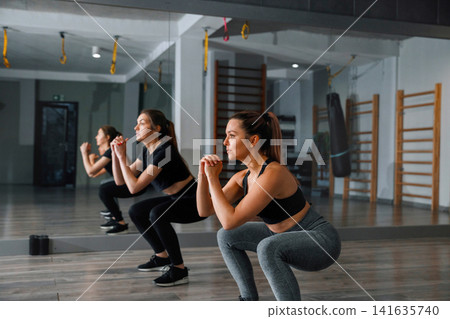 Women perform squats in a gym with wooden floors and exercise equipment during a workout session in the morning hours 141635740