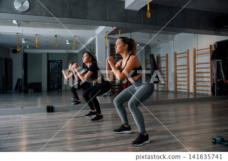 Women performing squats in a gym during a workout session in the morning with mirrors and fitness equipment in the background 141635741