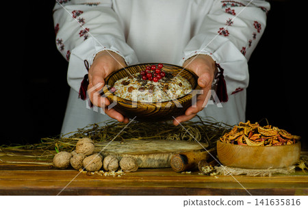 Woman in ukrainian native dresses standing behind table with kutia. Woman in national costume preparing kutya food for Christmas. Woman in ukrainian native dresses standing behind table with kutia. Woman in national costume preparing kutya food for Christmas. 141635816