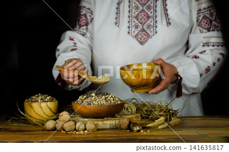 Woman in ukrainian native dresses standing behind table with kutia. Woman in national costume preparing kutya food for Christmas. Woman in ukrainian native dresses standing behind table with kutia. Woman in national costume preparing kutya food for Christmas. 141635817