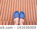 Top down view of feet wearing blue rubber sandals on a teak deck with black lines. Relaxing vacation vibe on a boat or luxury yacht. Concept of leisure, summer travel, and outdoor relaxation. 141636009