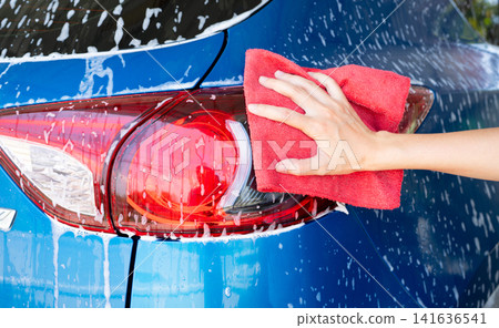 Car wash service. Car covered with white soap foam. Man hand holding red microfiber cloth and polish tail light of blue car. Auto care service business concept. Man cleaning and detailing luxury car. Car wash service. Car covered with white soap foam. Man hand holding red microfiber cloth and polish tail light of blue car. Auto care service business concept. Man cleaning and detailing luxury car. 141636541