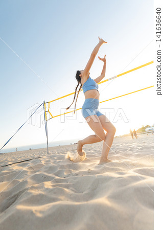 Woman playing beach volleyball during sunset near a volleyball net at the shore with sand all around her Woman playing beach volleyball during sunset near a volleyball net at the shore with sand all around her 141636640