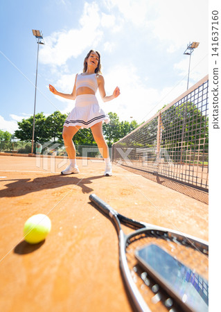 Young woman plays tennis on a sunny day at a sports park with a court and trees in the background during the afternoon Young woman plays tennis on a sunny day at a sports park with a court and trees in the background during the afternoon 141637160