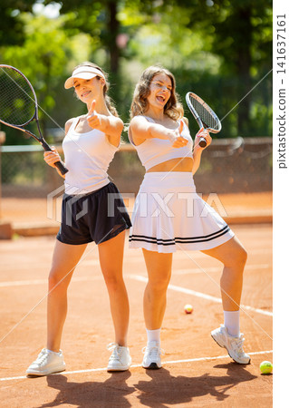 Two women pose with tennis rackets on a sunny day at a tennis court in summer Two women pose with tennis rackets on a sunny day at a tennis court in summer 141637161