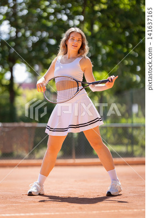 Young woman prepares to hit a tennis ball on a red clay court during a sunny day in a sports facility Young woman prepares to hit a tennis ball on a red clay court during a sunny day in a sports facility 141637165