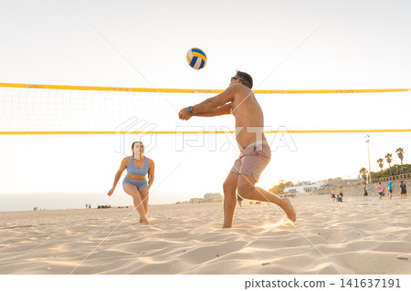 Players enjoy a game of beach volleyball at sunset along the sandy shore with spectators in the background Players enjoy a game of beach volleyball at sunset along the sandy shore with spectators in the background 141637191