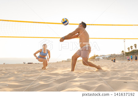 People play beach volleyball at sunset on a sandy beach with spectators in the background People play beach volleyball at sunset on a sandy beach with spectators in the background 141637192