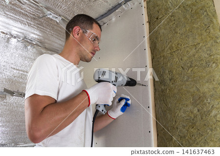Worker in goggles with screwdriver working on insulation. Drywall on wall beams, insulating rock wool staff in wooden frame. Comfortable warm home, economy, construction and renovation concept. Worker in goggles with screwdriver working on insulation. Drywall on wall beams, insulating rock wool staff in wooden frame. Comfortable warm home, economy, construction and renovation concept. 141637463