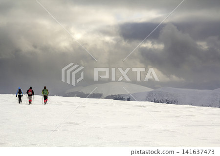 Winter mountain landscape. Three travelers tourist hikers in bright clothing with backpacks on snowy field walking towards distant mountain on cloudy dark blue stormy sky copy space background. Winter mountain landscape. Three travelers tourist hikers in bright clothing with backpacks on snowy field walking towards distant mountain on cloudy dark blue stormy sky copy space background. 141637473
