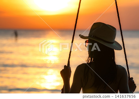 Silhouette of woman enjoying spectacular sunset from a beach swing overlooking the tranquil sea. Tropical vacation, luxury resort destination, mindfulness, coastal travel and summer lifestyle. Silhouette of woman enjoying spectacular sunset from a beach swing overlooking the tranquil sea. Tropical vacation, luxury resort destination, mindfulness, coastal travel and summer lifestyle. 141637656