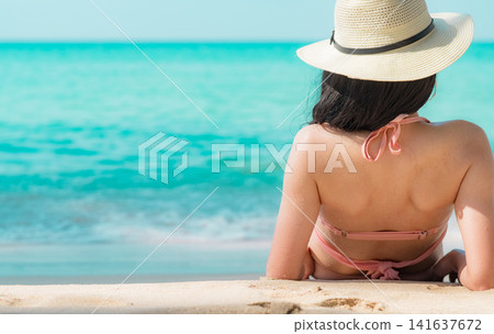 Rear view of woman relaxing on a tropical sandy beach while looking at the turquoise ocean, peaceful summer vacation, luxury travel destination and wellness lifestyle concept. Luxury beach resort. Rear view of woman relaxing on a tropical sandy beach while looking at the turquoise ocean, peaceful summer vacation, luxury travel destination and wellness lifestyle concept. Luxury beach resort. 141637672
