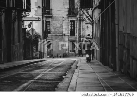 Municipal sanitation worker walking through quiet historic European street with leaf blower during morning maintenance, black and white urban documentary scene. Public service and city cleanliness. Municipal sanitation worker walking through quiet historic European street with leaf blower during morning maintenance, black and white urban documentary scene. Public service and city cleanliness. 141637769