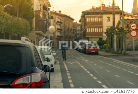 Cyclist riding along dedicated bicycle lane on quiet urban street with parked cars in residential neighborhood during morning light. Sustainable transportation, commuting and modern city mobility. Cyclist riding along dedicated bicycle lane on quiet urban street with parked cars in residential neighborhood during morning light. Sustainable transportation, commuting and modern city mobility. 141637776